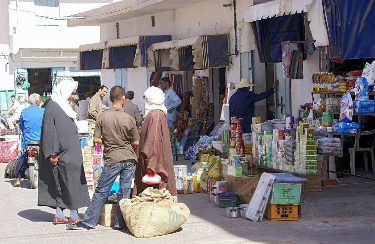 Passants et commerçants dans le souk de Tataouine, avec des étals de produits ménagers et de consommation exposés dans une rue commerçante animée.